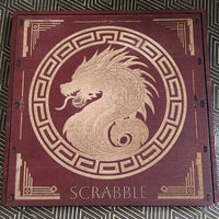 A square wooden Scrabble board with a dark reddish-brown finish and gold laser-engraved details, including a central dragon motif and the word 'SCRABBLE' at the bottom, placed on a patterned black and gold surface.