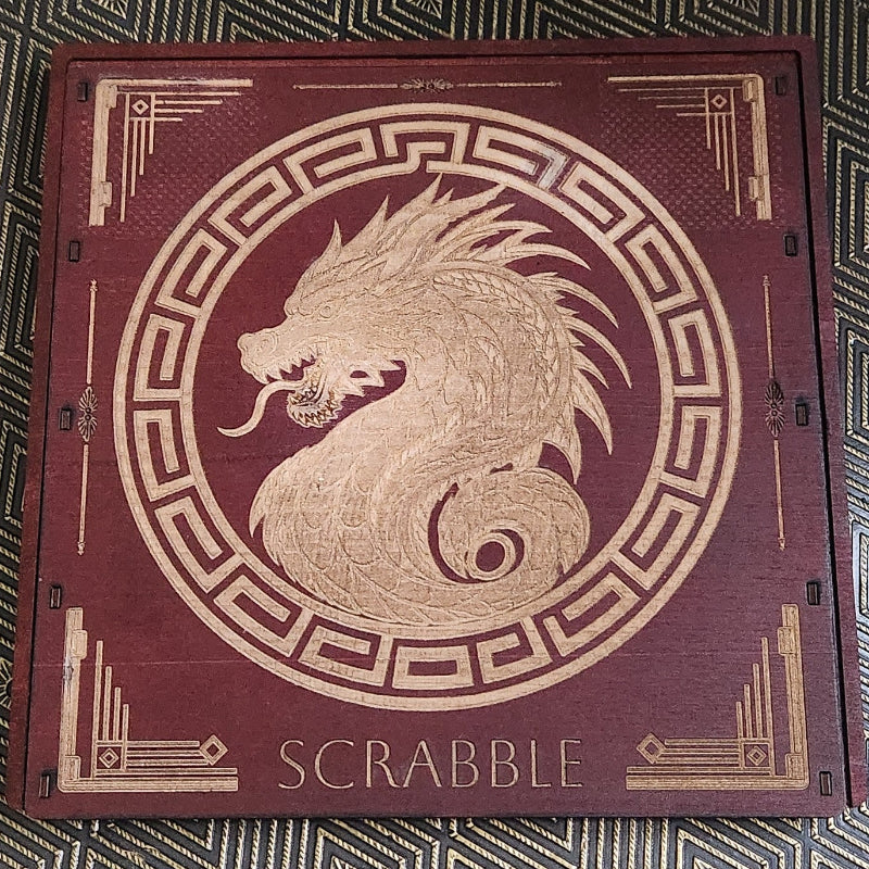 A square wooden Scrabble board with a dark reddish-brown finish and gold laser-engraved details, including a central dragon motif and the word 'SCRABBLE' at the bottom, placed on a patterned black and gold surface.