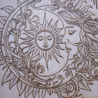 Close-up of a laser-engraved wood design featuring a crescent moon, a sun face, and a human profile surrounded by flowing hair and leaves.