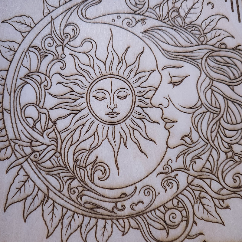 Close-up of a laser-engraved wood design featuring a crescent moon, a sun face, and a human profile surrounded by flowing hair and leaves.