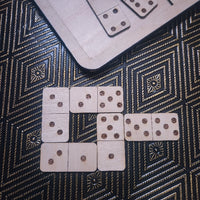 Wooden domino tiles arranged on a black and gold patterned surface, with additional dominoes partially visible on a wooden board in the top right.