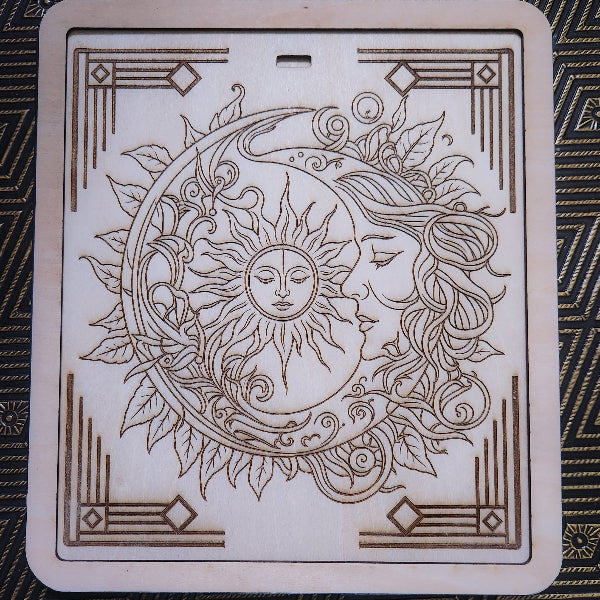 A square wooden decorative plaque or board featuring a laser-engraved sun and moon design with a woman's face, surrounded by leaves and ornate corner patterns, shown on a dark patterned fabric background.