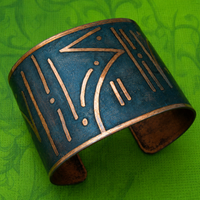 A wide cuff bracelet with a distressed teal-blue finish and engraved gold-toned geometric patterns, displayed on a textured green background.