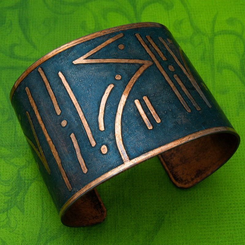 A wide cuff bracelet with a distressed teal-blue finish and engraved gold-toned geometric patterns, displayed on a textured green background.