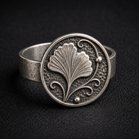 A silver ring with a circular face featuring an embossed ginkgo leaf design, photographed on a dark textured background.