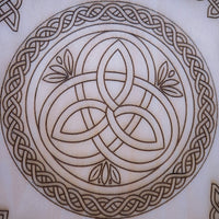 A close-up of a circular Celtic knot design etched or burned onto a light beige wood surface, showing a triquetra-style center surrounded by an ornate border.