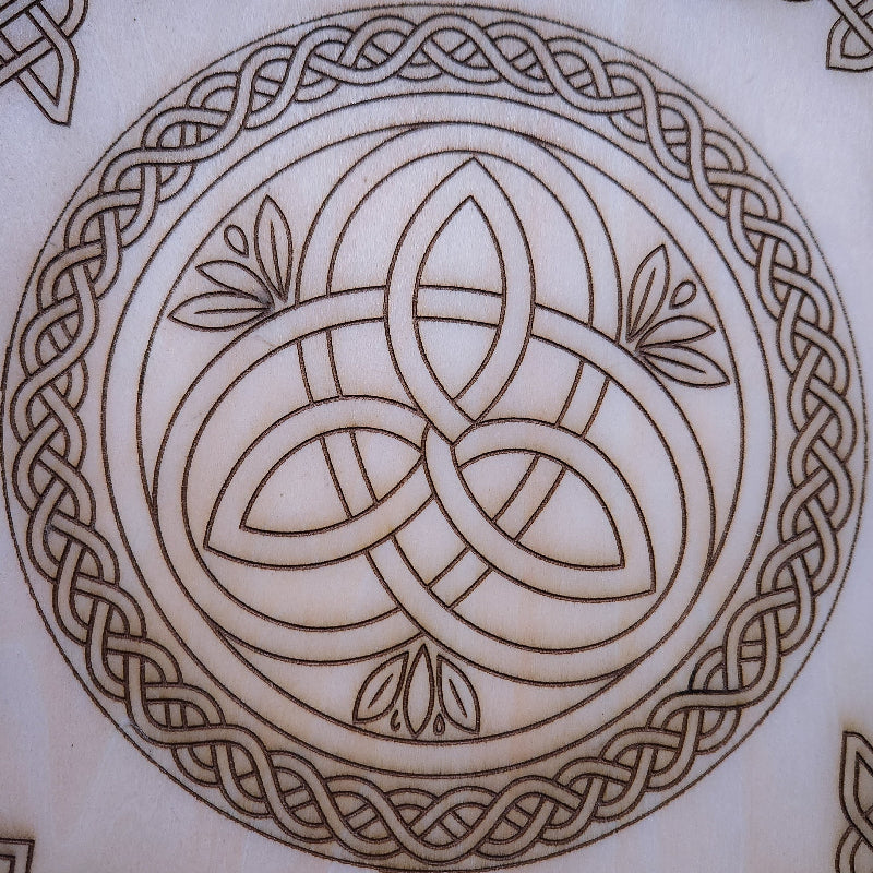 A close-up of a circular Celtic knot design etched or burned onto a light beige wood surface, showing a triquetra-style center surrounded by an ornate border.