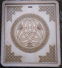 A square light wood plaque featuring Celtic knot designs, including a central triquetra symbol inside a circle and corner knot patterns, photographed flat on a dark patterned fabric background.