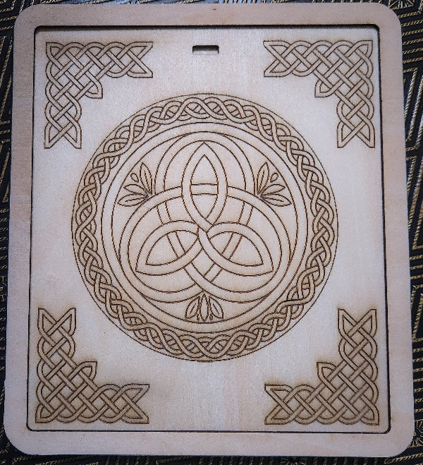 A square light wood plaque featuring Celtic knot designs, including a central triquetra symbol inside a circle and corner knot patterns, photographed flat on a dark patterned fabric background.