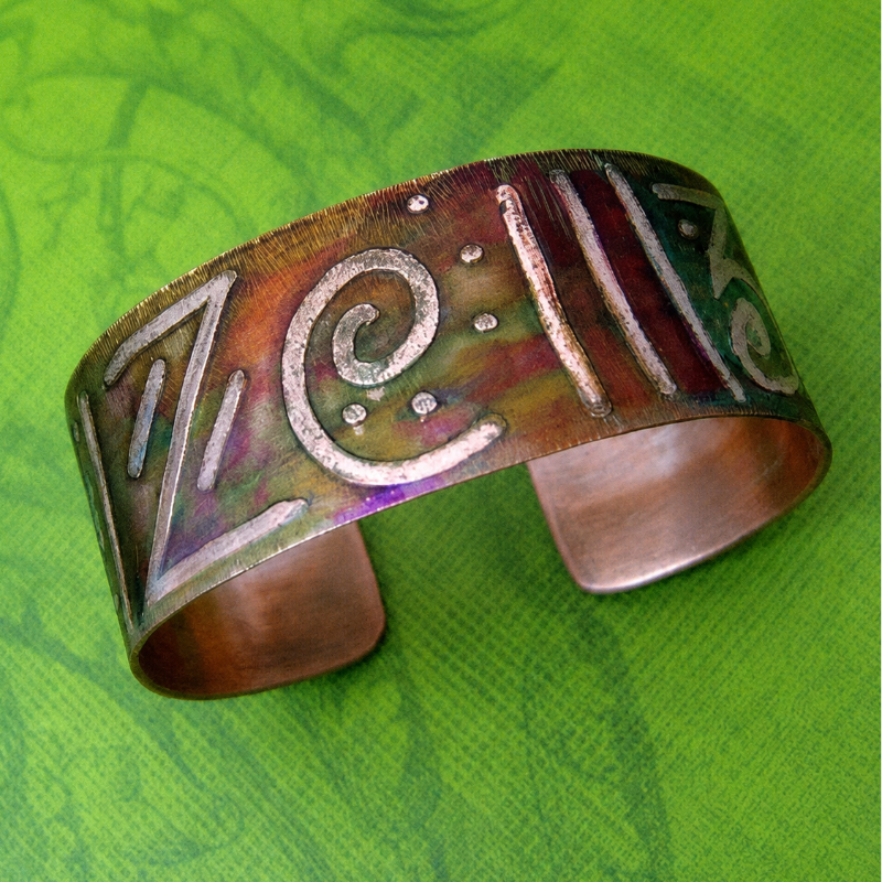 A handmade copper cuff bracelet with hand-painted purple, green, and gold accents, featuring stylized lettering, displayed on a textured green background.