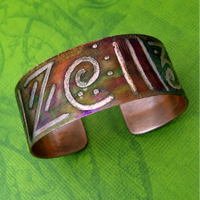 A handmade copper cuff bracelet with hand-painted purple, green, and gold accents, featuring stylized lettering, displayed on a textured green background.