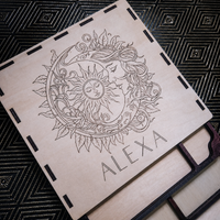 A square wooden plaque or coaster with a laser-engraved design of a sun and moon surrounded by leaves, with the name 'ALEXA' printed at the bottom. The piece is placed on a black patterned fabric background.