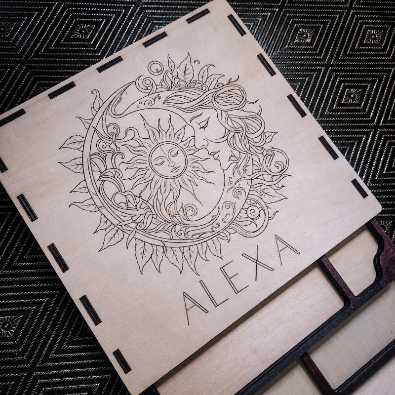 A square wooden plaque or coaster with a laser-engraved design of a sun and moon surrounded by leaves, with the name 'ALEXA' printed at the bottom. The piece is placed on a black patterned fabric background.