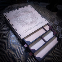 A wooden book organizer with a laser-engraved sun and moon design and the text 'STUDIO NAVARRI' on the cover, shown open with multiple card slots on a patterned dark table.