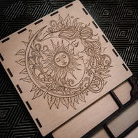 A square wooden board or tray with a laser-engraved celestial design featuring a sun and moon surrounded by leaves, placed on a dark patterned surface.