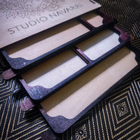 A close-up of a wooden multi-compartment organizer tray with dark stained edges and light natural wood interiors, shown on a patterned black and gold surface. A branded lid or insert reading STUDIO NAVARRI is visible at the top.