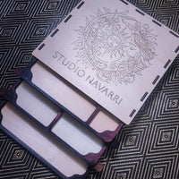 A wooden multi-compartment organizer tray with a hinged lid engraved with a sun and moon design and the text 'STUDIO NAVARRI', shown on a dark patterned fabric background.