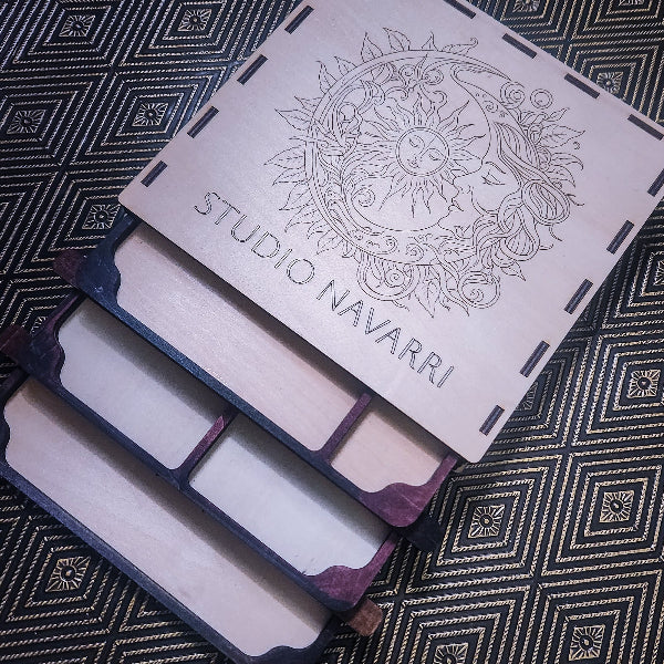 A wooden multi-compartment organizer tray with a hinged lid engraved with a sun and moon design and the text 'STUDIO NAVARRI', shown on a dark patterned fabric background.