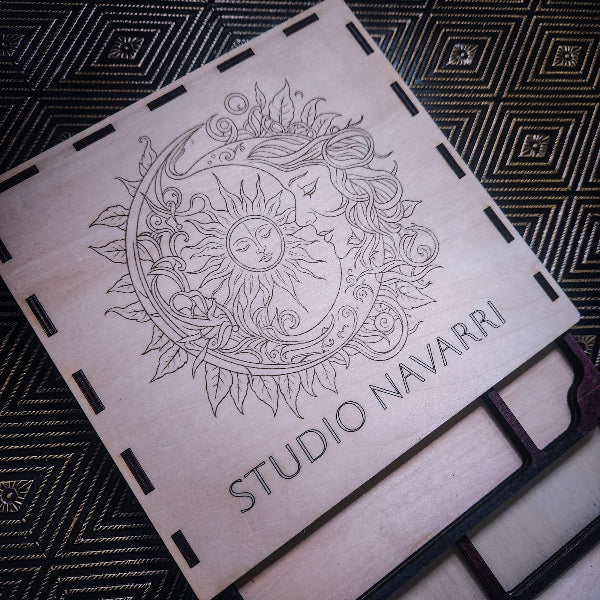 A square wooden box or plaque with laser-engraved artwork of a sun and moon surrounded by leaves, with the text 'STUDIO NAVARRI' printed on the front, placed on a dark patterned fabric background.