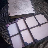 A wooden multi-compartment organizer tray with a matching engraved wooden box lid featuring a sun design and the text 'STUDIO NAVARRI', placed on a patterned black and gold surface.