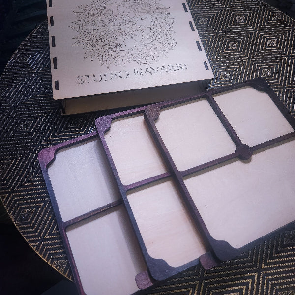 A wooden multi-compartment organizer tray with a matching engraved wooden box lid featuring a sun design and the text 'STUDIO NAVARRI', placed on a patterned black and gold surface.