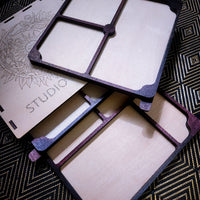 Laser-cut wooden jewelry display stands with a dark purple painted border, shown in an overlapping stack on a black and gold patterned rug. One piece features an engraved floral design and the word 'STUDIO'.