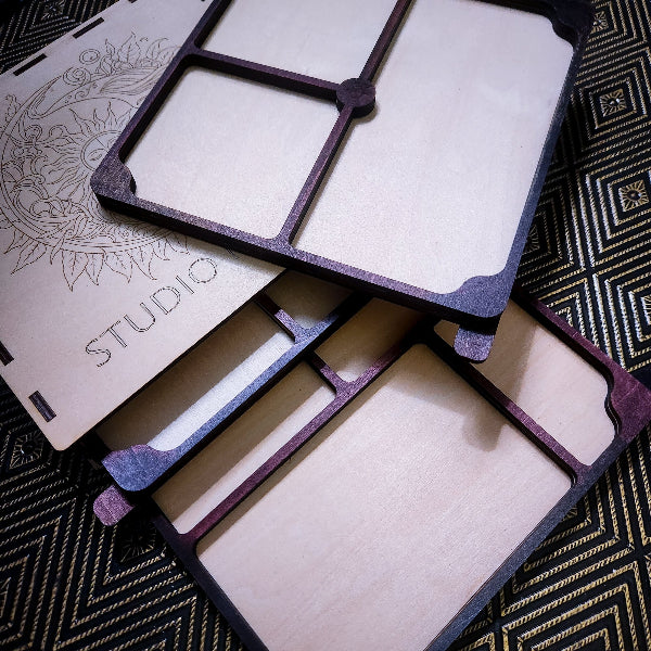 Laser-cut wooden jewelry display stands with a dark purple painted border, shown in an overlapping stack on a black and gold patterned rug. One piece features an engraved floral design and the word 'STUDIO'.