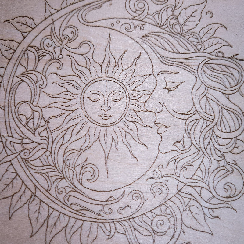 A close-up of a light beige wood with an intricate engraved line-art design featuring a sun face, moon face, flowing hair, and leafy ornamental swirls.