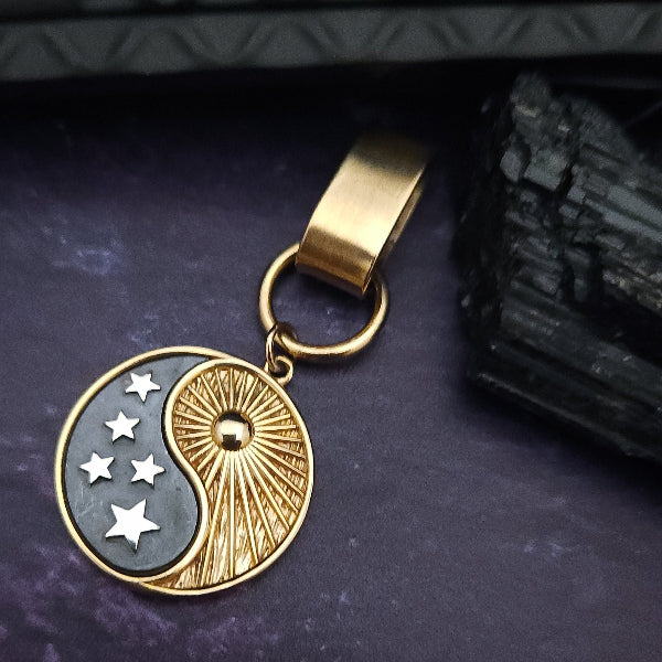 A gold and silver-toned yin-yang keychain charm with stars on a dark purple textured background next to a black crystal.