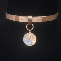 A gold-toned mesh choker necklace with a circular pendant featuring a split yin-yang design, one half in gold with sunburst lines and stars, and the other half in light blue with moon phases.