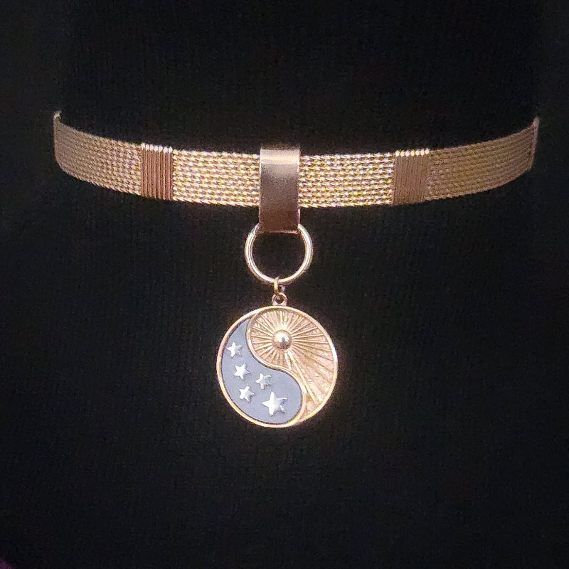 A gold-toned mesh choker necklace with a circular pendant featuring a split yin-yang design, one half in gold with sunburst lines and stars, and the other half in light blue with moon phases.