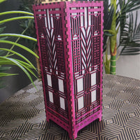 Pink decorative lamp with geometric patterns on a patterned surface with plants in the background