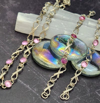 A close-up of a handmade necklace featuring iridescent oval cabochon stones, twisted gold-tone chain links, and small pink gemstone accents, displayed on a dark textured surface with a white crystal slab behind it.