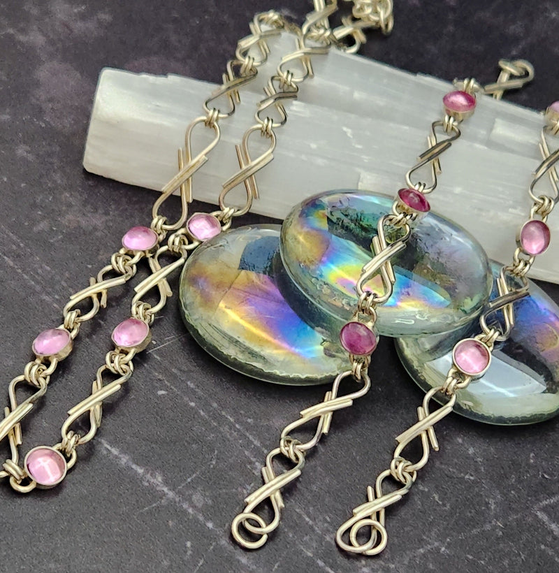 A close-up of a handmade necklace featuring iridescent oval cabochon stones, twisted gold-tone chain links, and small pink gemstone accents, displayed on a dark textured surface with a white crystal slab behind it.