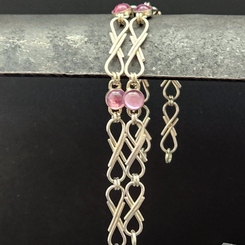 A sterling ribbon link bracelet with pink round gemstone accents, displayed against a black background with a textured gray bar at the top.