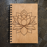 A spiral-bound notebook with a light wood cover featuring a laser-engraved lotus flower design, placed flat on a black and gold patterned surface.
