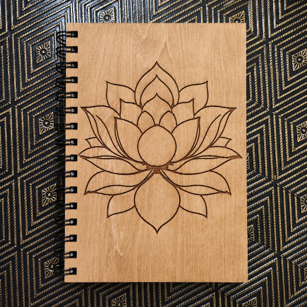 A spiral-bound notebook with a light wood cover featuring a laser-engraved lotus flower design, placed flat on a black and gold patterned surface.
