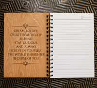 An open spiral-bound notebook with a natural wood cover on the left page and lined white pages on the right, placed on a dark patterned surface. The cover features engraved inspirational text and small lotus symbols.