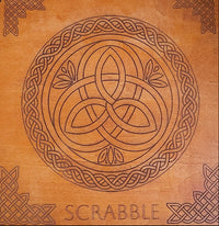 A square wooden board or plaque featuring a carved Celtic knot design in the center, surrounded by a circular braided border and the word 'SCRABBLE' at the bottom, with decorative corner motifs on a warm wood background.