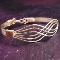 A multi-strand wire wrap bracelet featuring alternating silver and gold-toned wires, with wraps at the ends, displayed on a dark purple textured background.