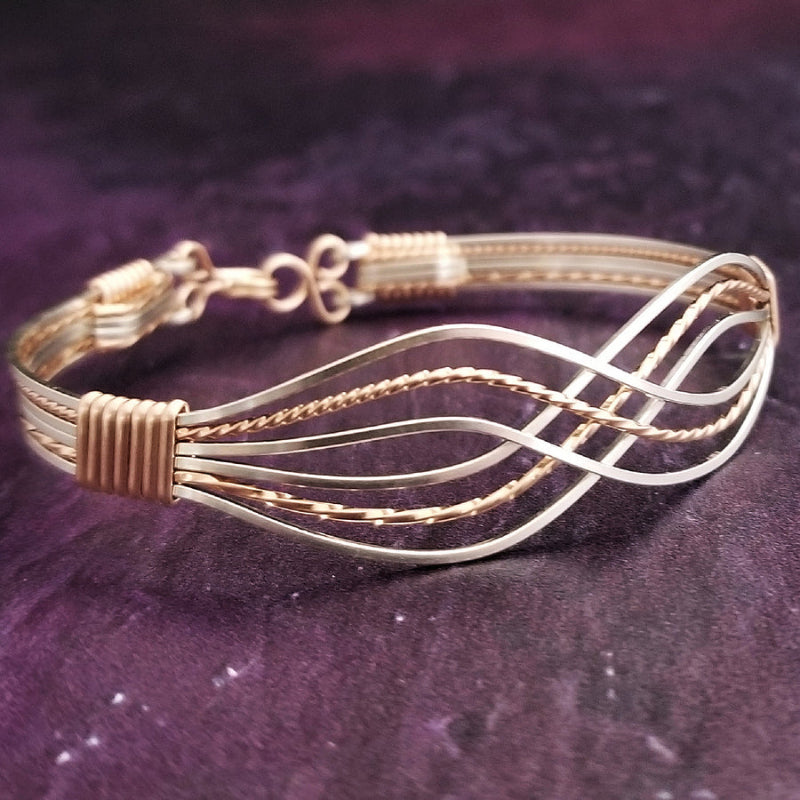 A multi-strand wire wrap bracelet featuring alternating silver and gold-toned wires, with wraps at the ends, displayed on a dark purple textured background.