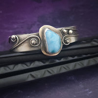 This unique piece features a breathtaking Larimar cabochon and two faceted Amethyst stones, adorned with intricate handcrafted waves. The gemstone is carefully set in a handcrafted sterling silver bezel, making it a true one-of-a-kind design. All Sterling Silver.
