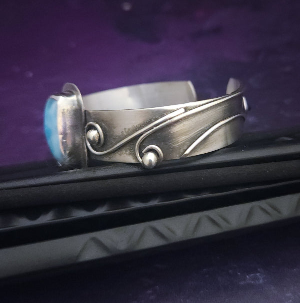 This unique piece features a breathtaking Larimar cabochon and two faceted Amethyst stones, adorned with intricate handcrafted waves. The gemstone is carefully set in a handcrafted sterling silver bezel, making it a true one-of-a-kind design. All Sterling Silver.