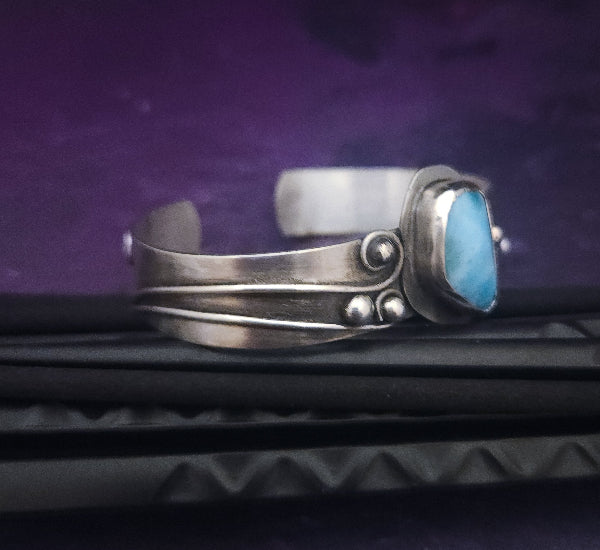 This unique piece features a breathtaking Larimar cabochon and two faceted Amethyst stones, adorned with intricate handcrafted waves. The gemstone is carefully set in a handcrafted sterling silver bezel, making it a true one-of-a-kind design. All Sterling Silver.