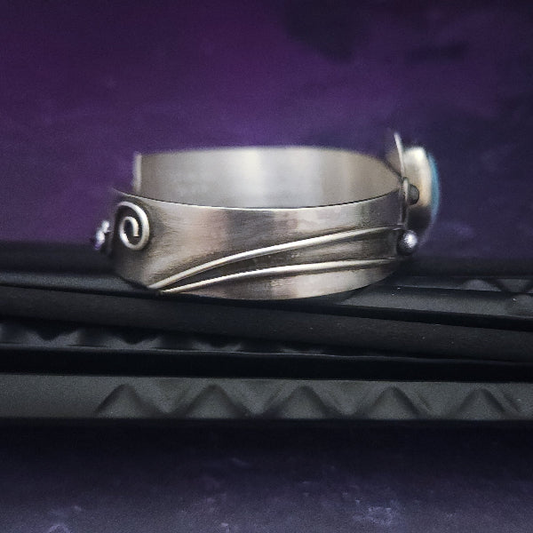 This unique piece features a breathtaking Larimar cabochon and two faceted Amethyst stones, adorned with intricate handcrafted waves. The gemstone is carefully set in a handcrafted sterling silver bezel, making it a true one-of-a-kind design. All Sterling Silver.