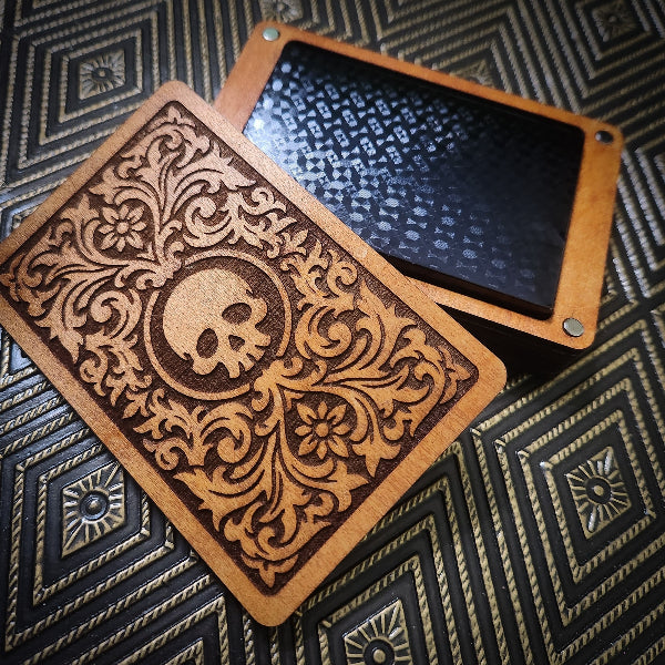 A wooden box with an ornate laser-engraved lid featuring a skull and floral scrollwork, shown next to an open lid revealing a black textured interior, set on a dark patterned surface.