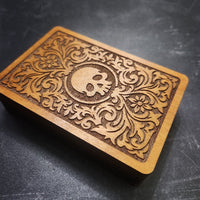 A rectangular wooden box with a carved floral scroll pattern and a skull emblem in the center, photographed on a dark textured surface.
