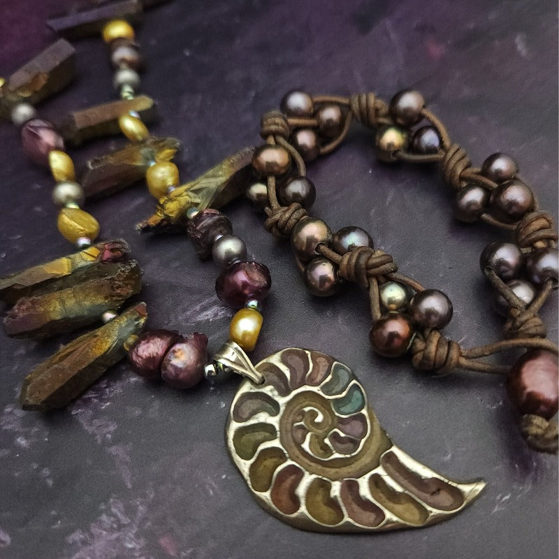 Make a bold statement with MARNIA, the one-of-a-kind necklace featuring titanium druzy, freshwater pearls, and a handcrafted bronze medallion ammonite. Turn heads with the vibrant colors and asymmetrical design, guaranteed to make you look and feel unforgettable!