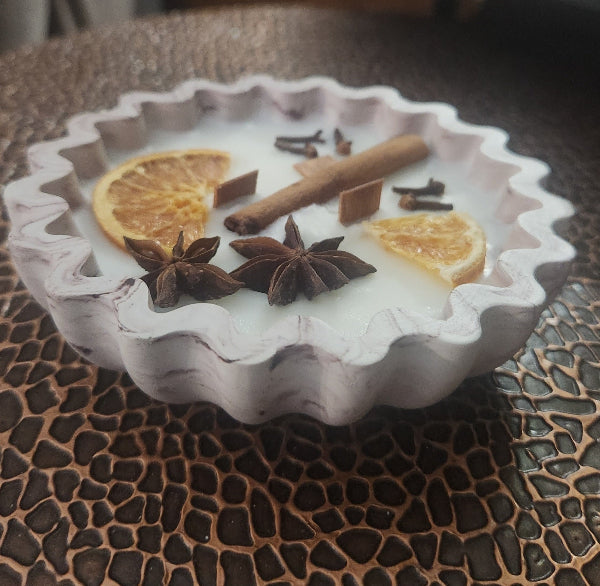 Candle in a decorative bowl with star anise, cinnamon sticks, and orange slices on a textured surface