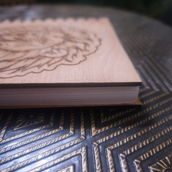 A close-up of a light natural wood notebook or journal cover with a laser-engraved floral or wing-like design, resting on a dark patterned surface.